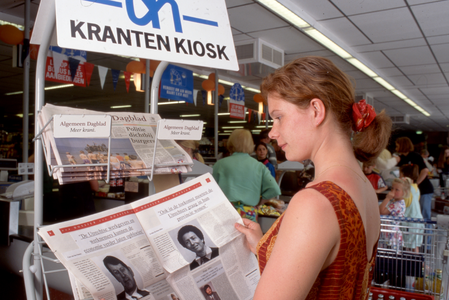 846316 Afbeelding van een jonge vrouw die de krant Het Utrechts Nieuws doorbladert bij het krantenrek van supermarkt ...